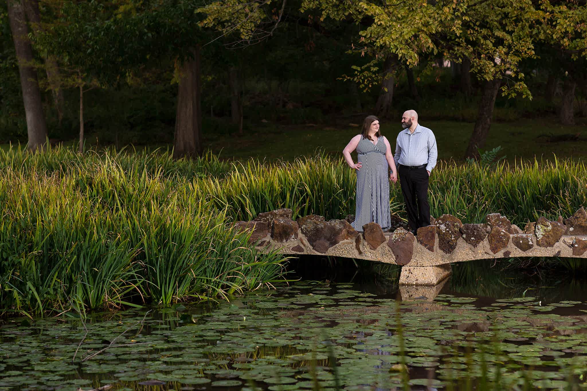 Engagement photo at Texas Women's University with engaged couple standing together looking at each other on a bridge over a lake filled with Lilly pads with trees surrounding them