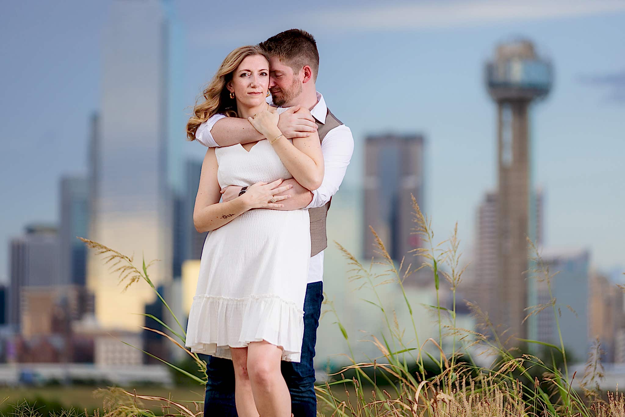 Man nuzzling into his fiancee's neck during outdoor engagement session wearing white dress with downtown Dallas in the background